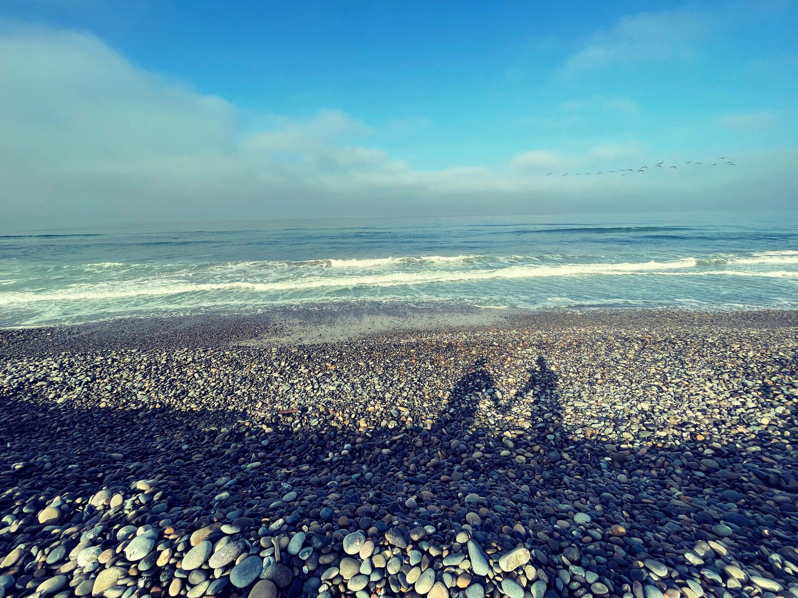 Two long shadows thrown across a pebble beach, surf breaking beyond