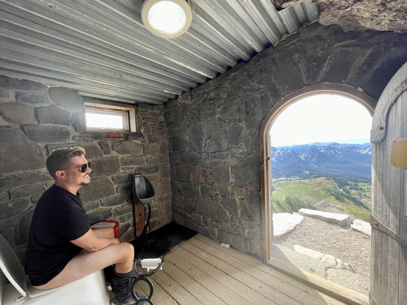 Jesse sitting on the trailside toilet at Mount Rainier, the door open to a mountain view